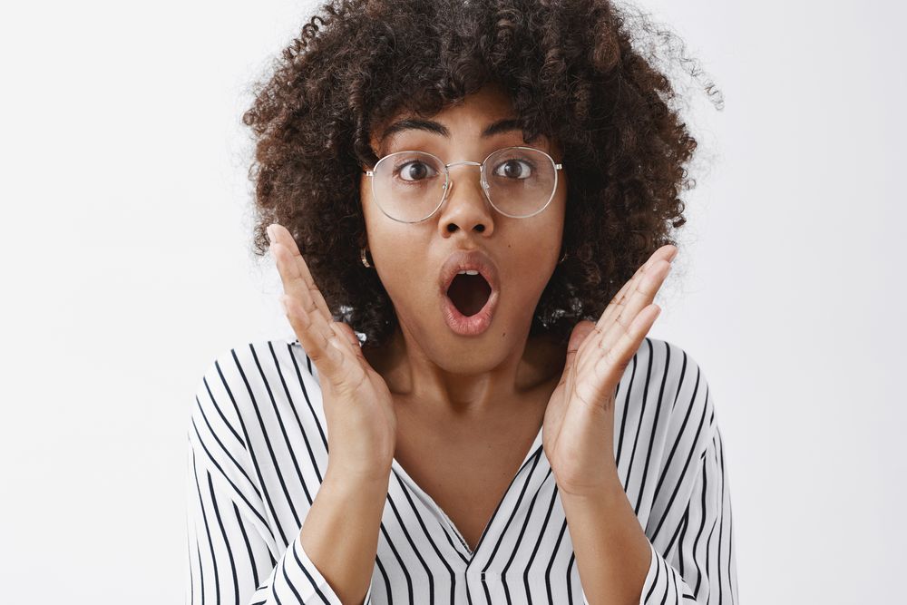 Headshot of impressed attractive african american female with afro hairstyle in transparent glasses gasping opening mouth from amazement and shock holding hands near face standing shook over gray wall