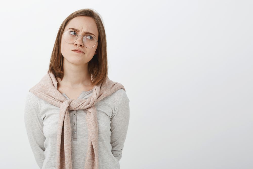 Portrait of perplexed gloomy smart girlfriend in glasses with short brown hair turning away looking right frowning and pursing lips being concerned and thoughtful solving dilemma over grey wall