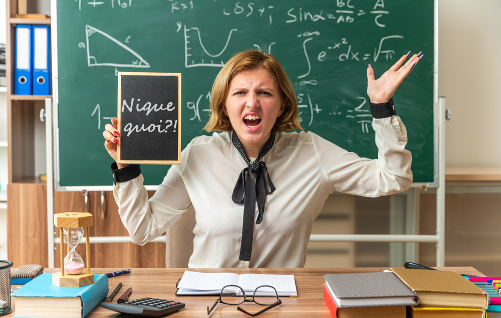 young female teacher sits at table with school tools holding mini blackboard in classroom