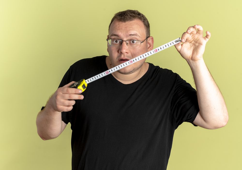 overweight man in glasses wearing black t-shirt holding ruler looking at it surprised standing over light background