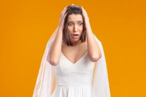 bride in beautiful wedding dress looking aside being shocked with hands on her head standing over orange background